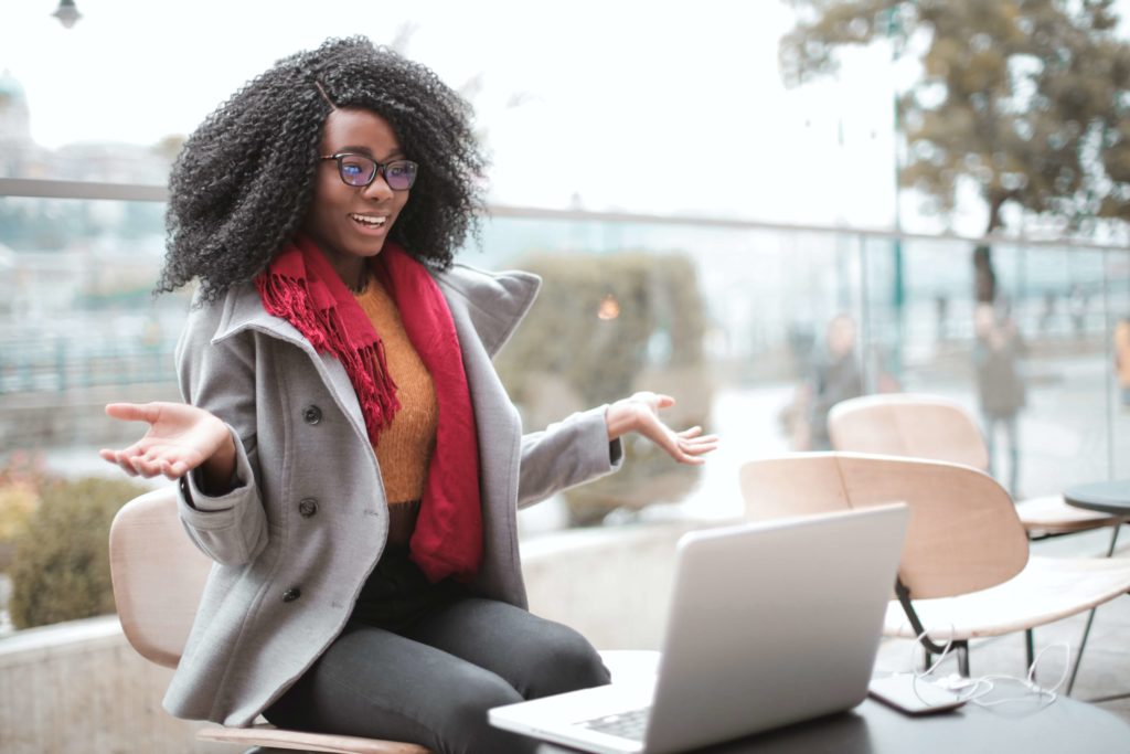 A young Black woman in a gray peacoat and red scarf sits outside a cafe with her laptop on a Zoom meeting with her nonprofit marketing team.
