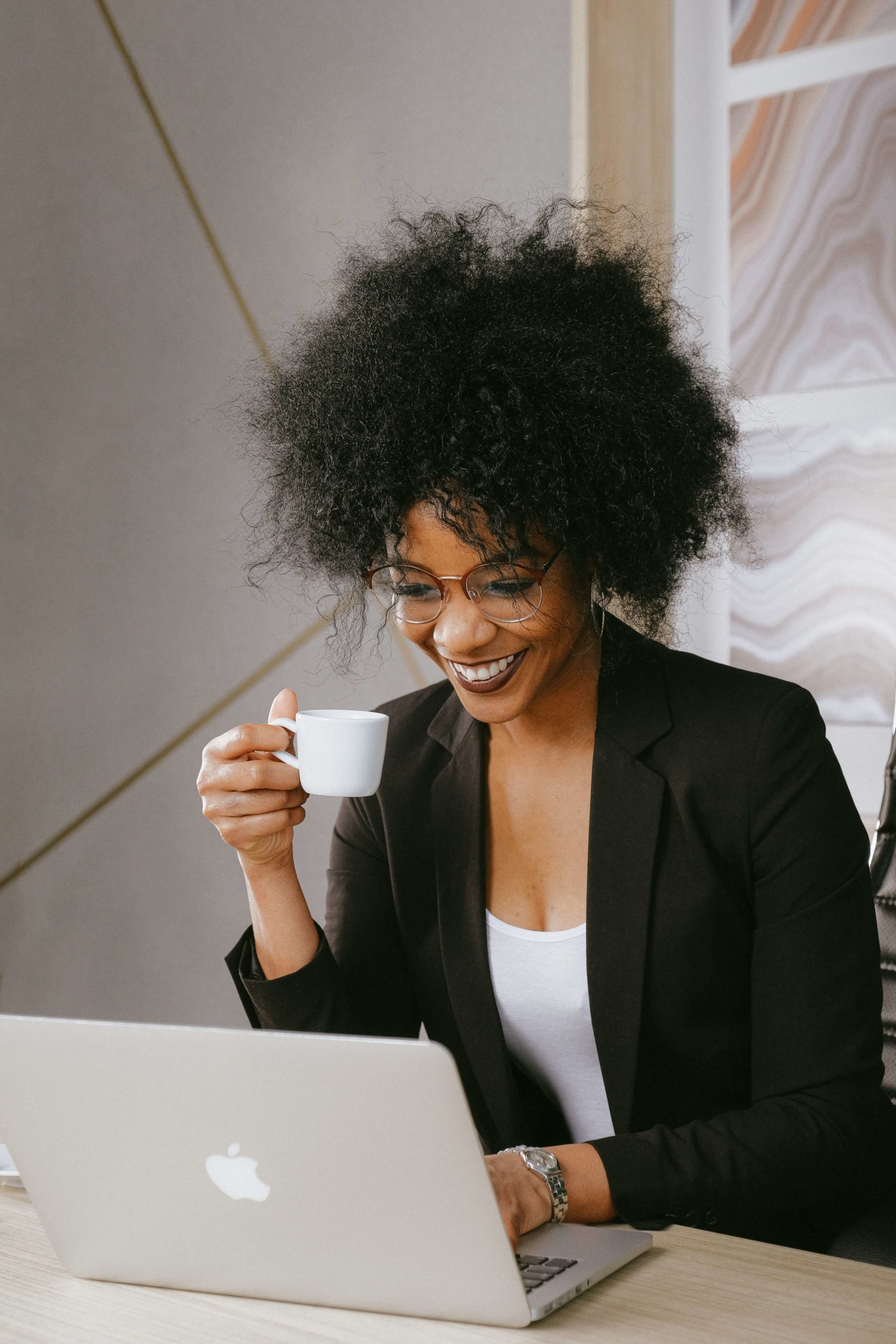 A young Black woman in a black blazer holds a small espresso mug while she uses her laptop to learn more about using nonprofit Facebook ads.