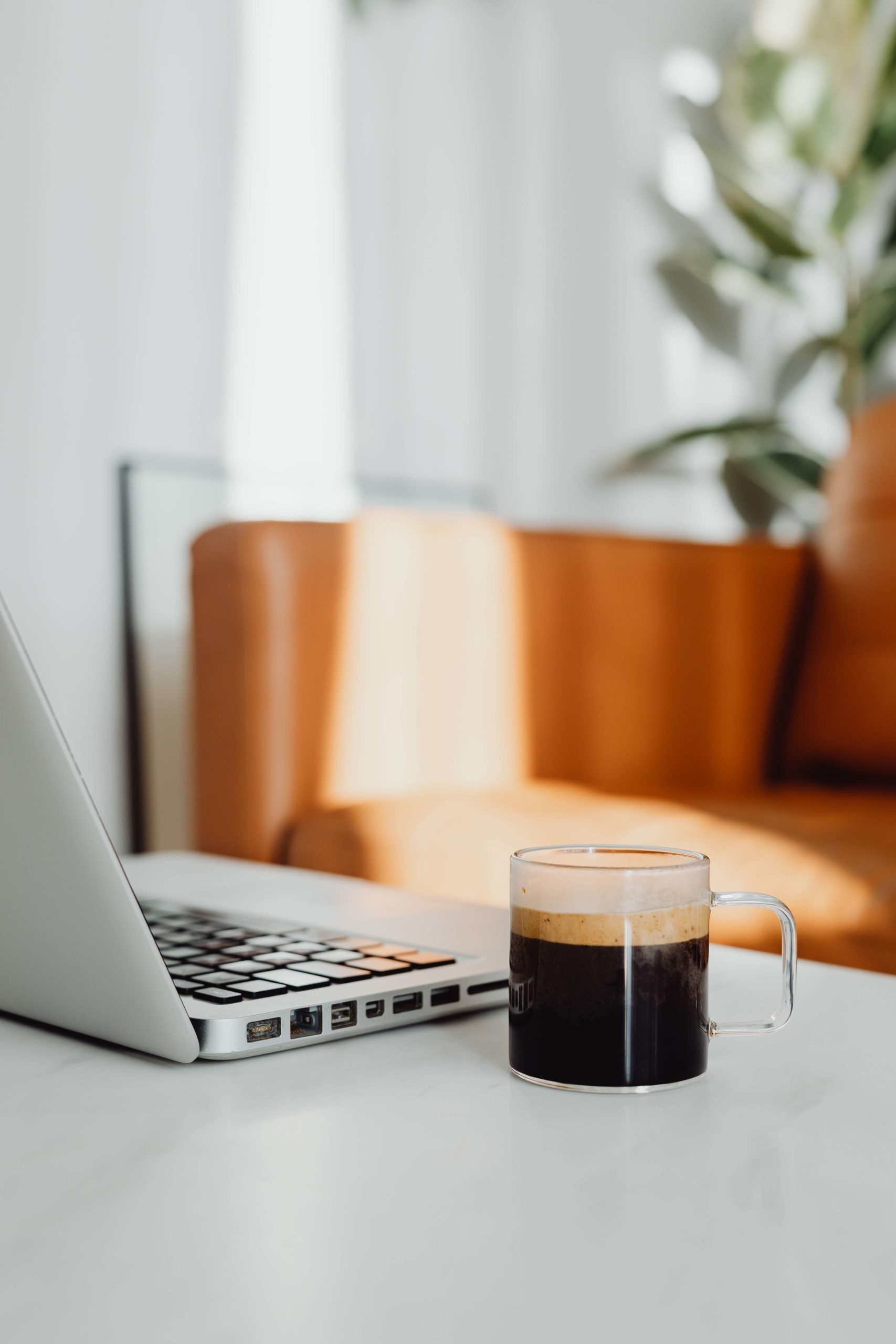 An americano in a glass mug sits next to a laptop on a white tabletop with a tan sofa in the background.